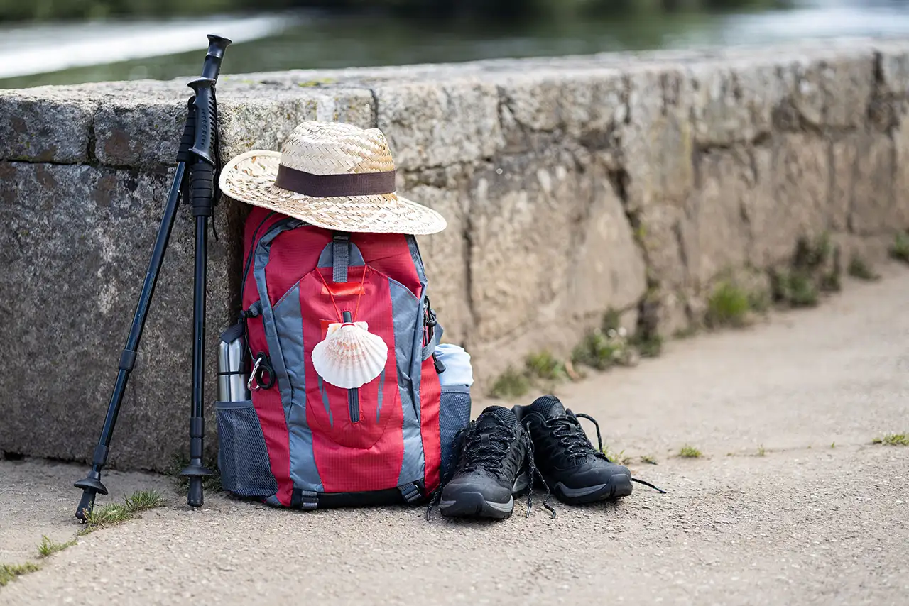 Mochila de peregrino con bastones, sombrero y botas apoyados junto a un muro de piedra del Camino de Santiago.