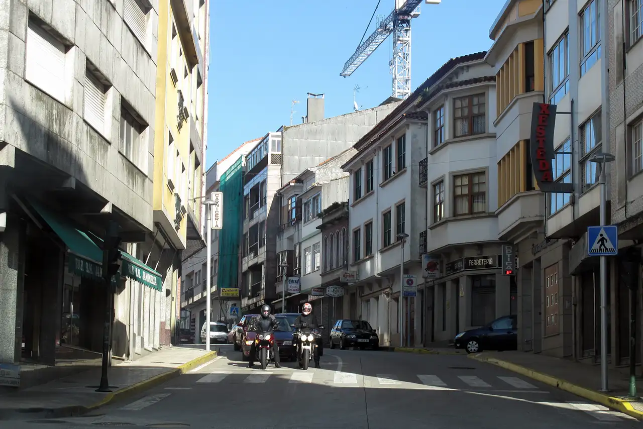 Calle comercial de Ordes con edificios de varias plantas, paso de peatones en primer plano y dos motoristas circulando por la vía.