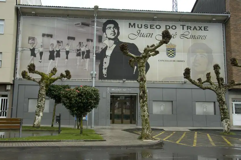 Fachada del Museo do Traxe Juan José Linares en Ordes, con el gran mural exterior, árboles podados delante del edificio y la entrada principal al centro.